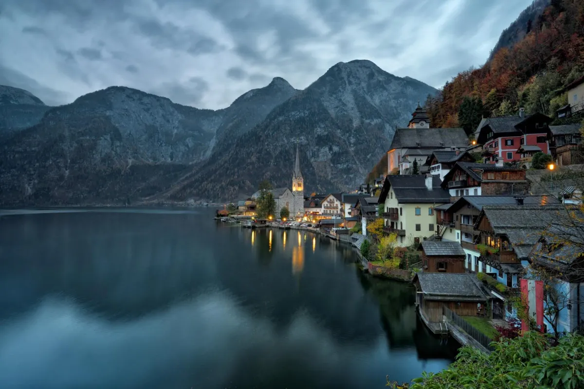 Hallstatt village in Austria at dusk, lakeside houses and a church tower illuminated with lights, reflected in the still lake water