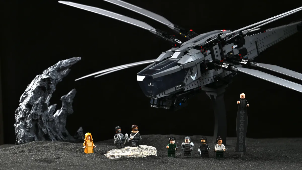 The LEGO Dune Ornithopter on its display stand with wings spread, photographed against black sand and rock formations with the minifigure crew gathered underneath