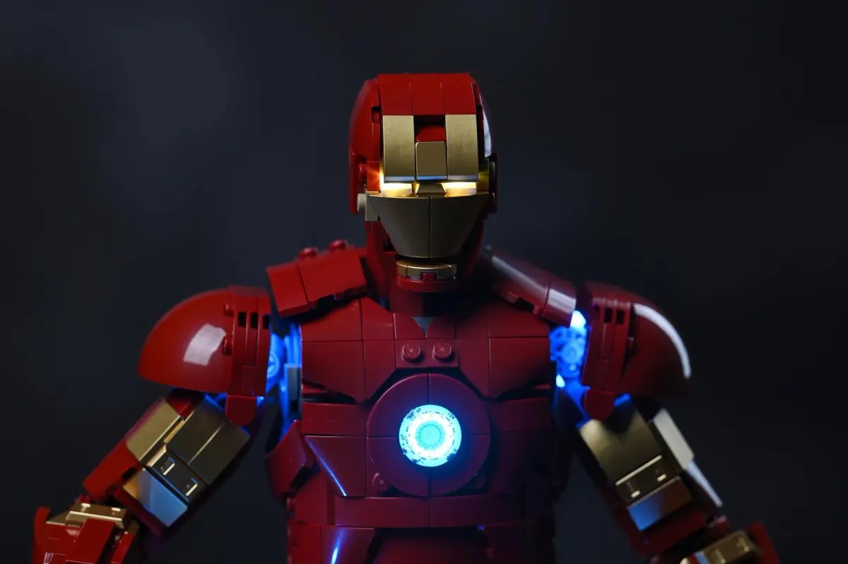 The upper body of the LEGO Iron Man Mark 3 in a dark room with the BriksMax kit installed, arc reactor and shoulder repulsors glowing blue and the eyes lit gold