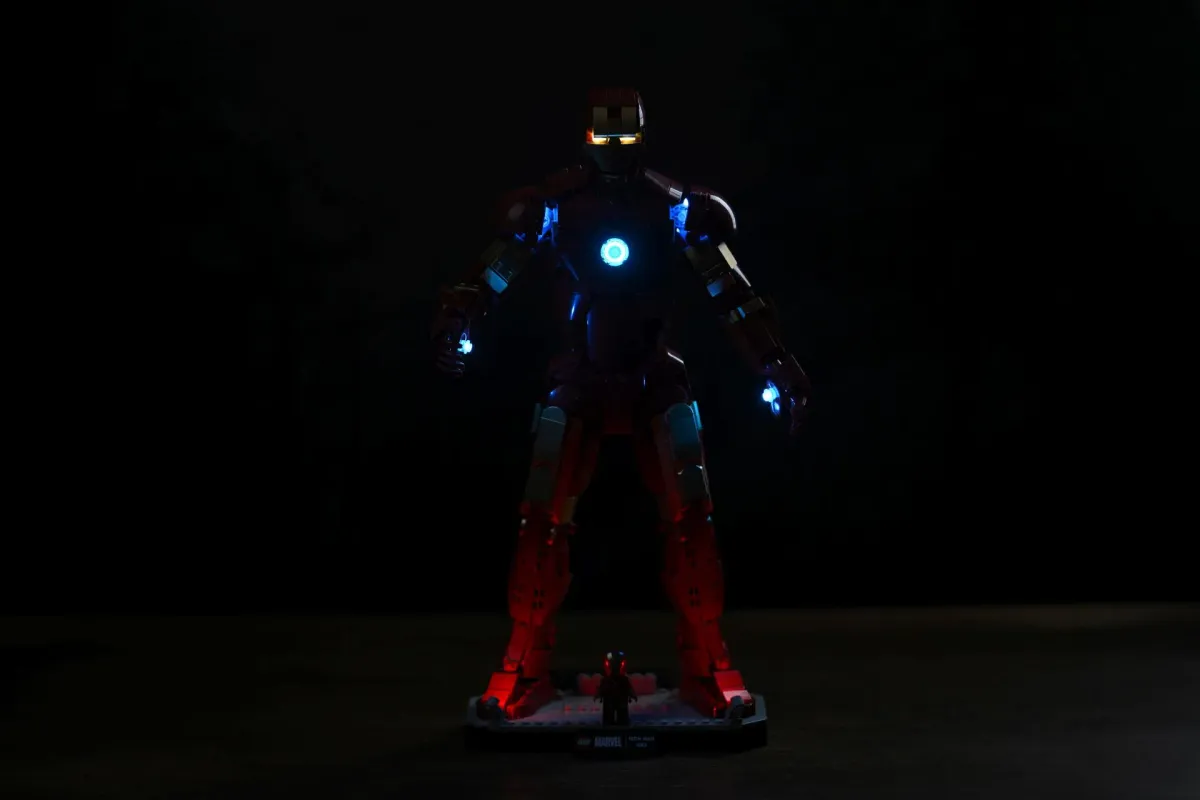 The LEGO Iron Man Mark 3 in near-total darkness with the BriksMax kit on, arc reactor and repulsors glowing and a red underglow illuminating the display base