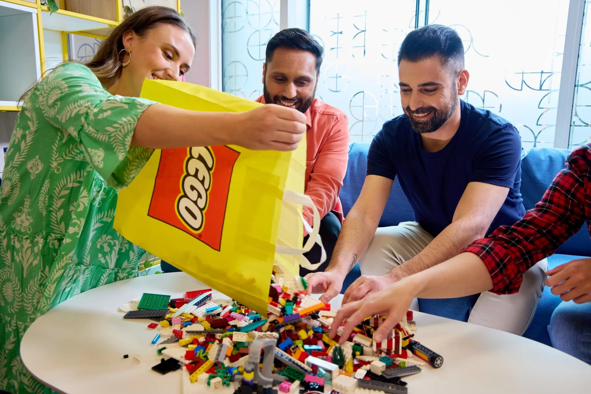 Three adults emptying a yellow LEGO bag onto a table full of bricks - official LEGO lifestyle photo