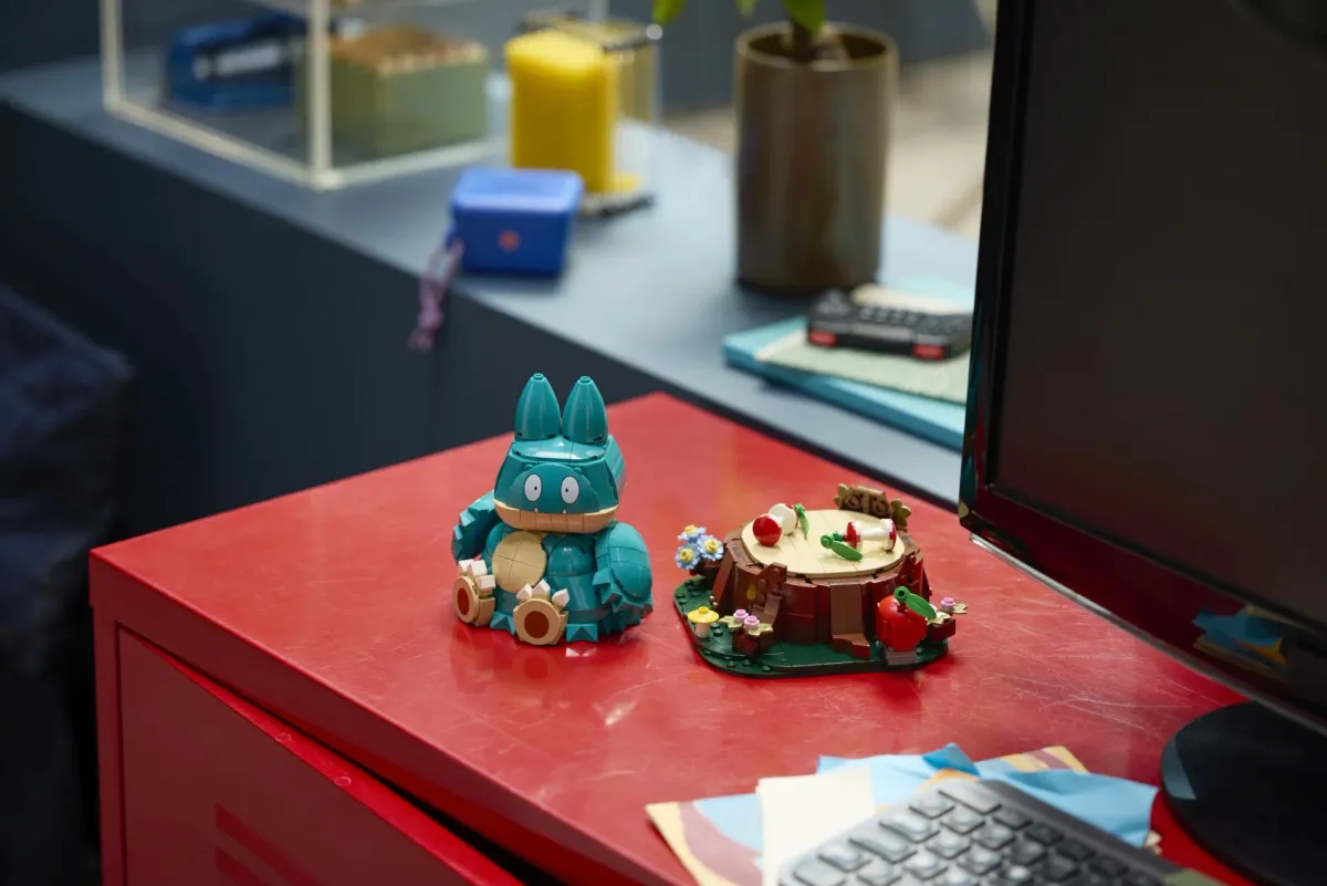 The LEGO Munchlax figure displayed next to its separate tree stump base with apple, mushrooms and flower details on a red desk