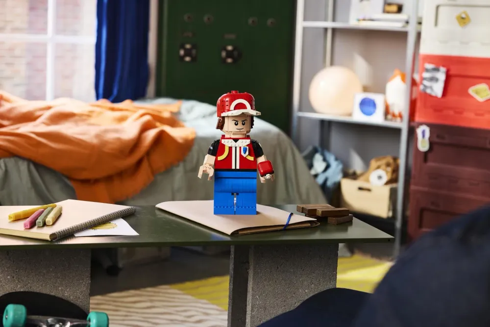 The LEGO Pokémon Up-Scaled Red Minifigure (40868) built and standing on a desk in a kid's room, wearing the red cap and vest and holding a Poké Ball