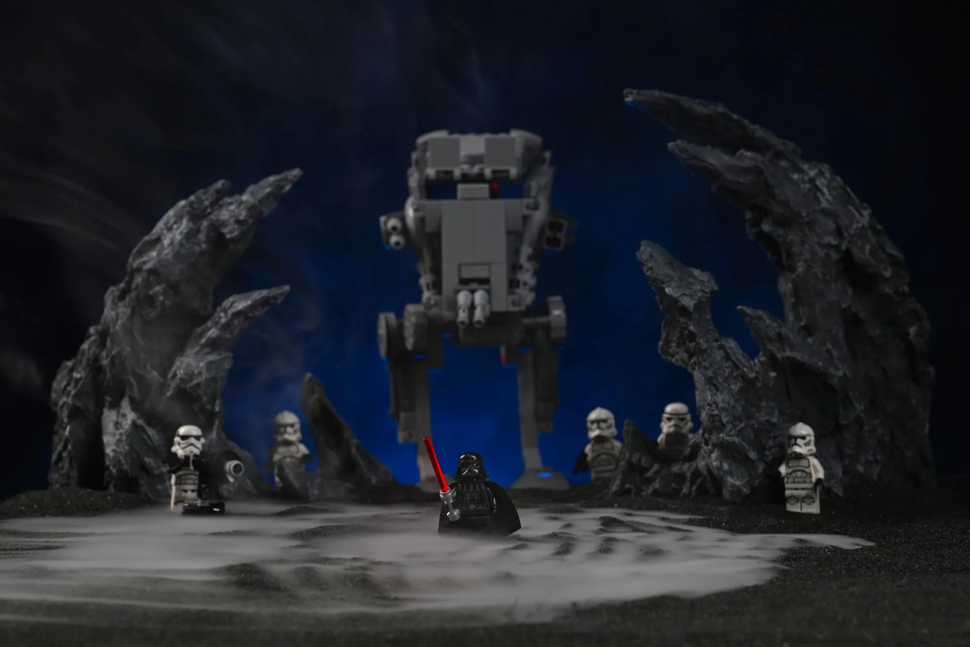 A LEGO Darth Vader minifigure standing in front of an AT-ST walker and a squad of clone troopers, with thick dry-ice-style fog pooling across the black ground and rising between dark rock formations