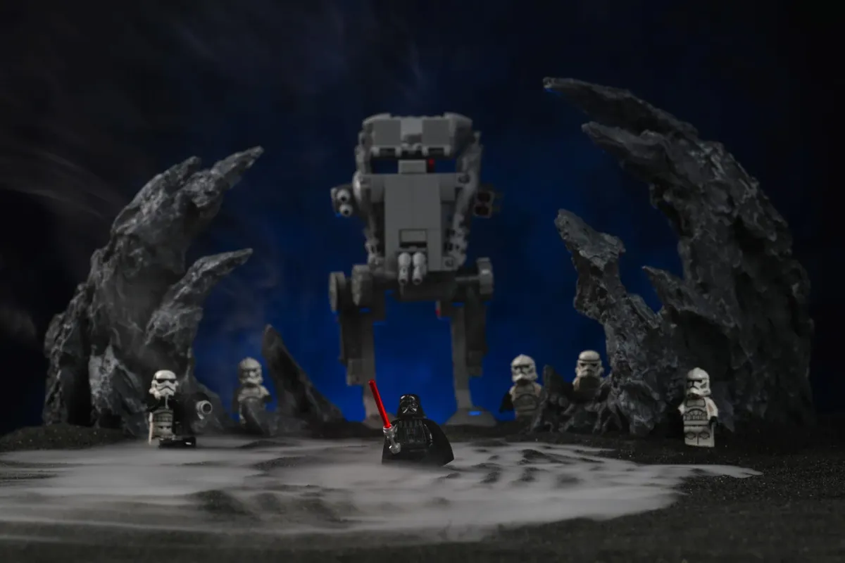 A LEGO Darth Vader minifigure standing in front of an AT-ST walker and a squad of clone troopers, with thick dry-ice-style fog pooling across the black ground and rising between dark rock formations