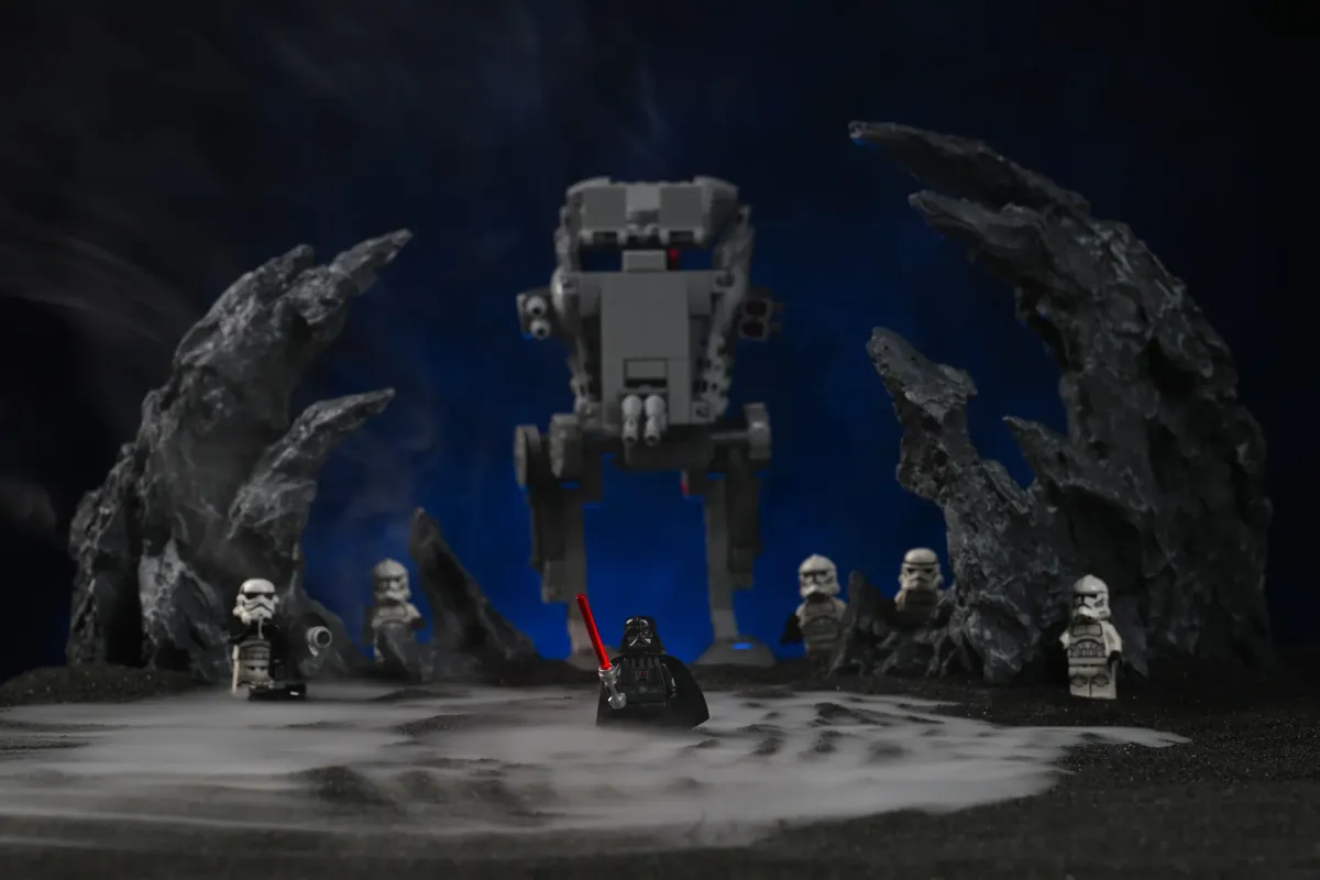 A LEGO Darth Vader minifigure standing in front of an AT-ST walker and a squad of clone troopers, with thick dry-ice-style fog pooling across the black ground and rising between dark rock formations