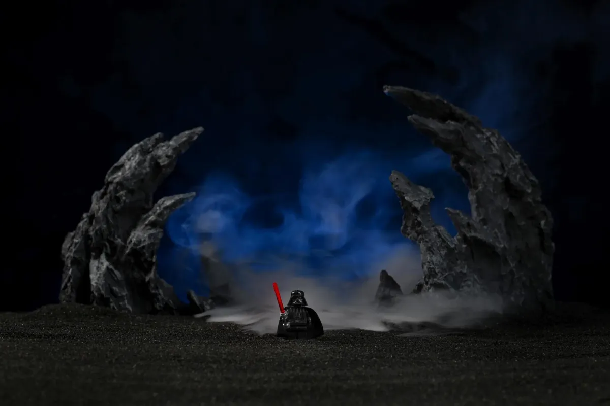 A lone LEGO Darth Vader minifigure with a lit red lightsaber, standing between two jagged black rock formations with blue-lit haze drifting through the background