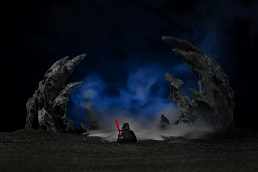 A lone LEGO Darth Vader minifigure with a lit red lightsaber, standing between two jagged black rock formations with blue-lit haze drifting through the background