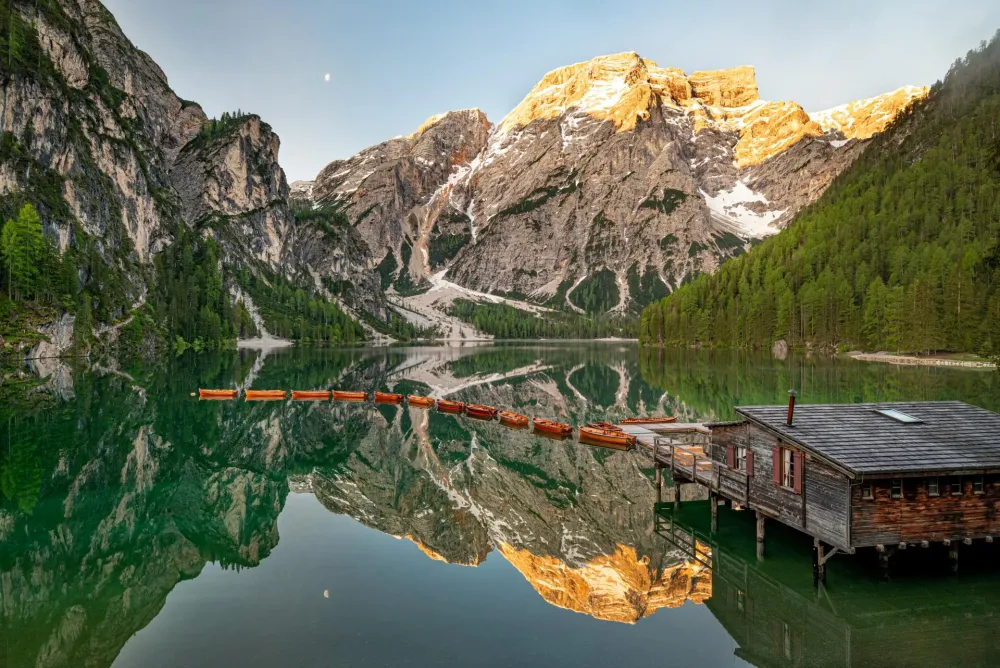 Row of wooden boats on the emerald water of Lake Pragser Wildsee in the Dolomites at sunrise, with the Seekofel peak glowing orange behind them