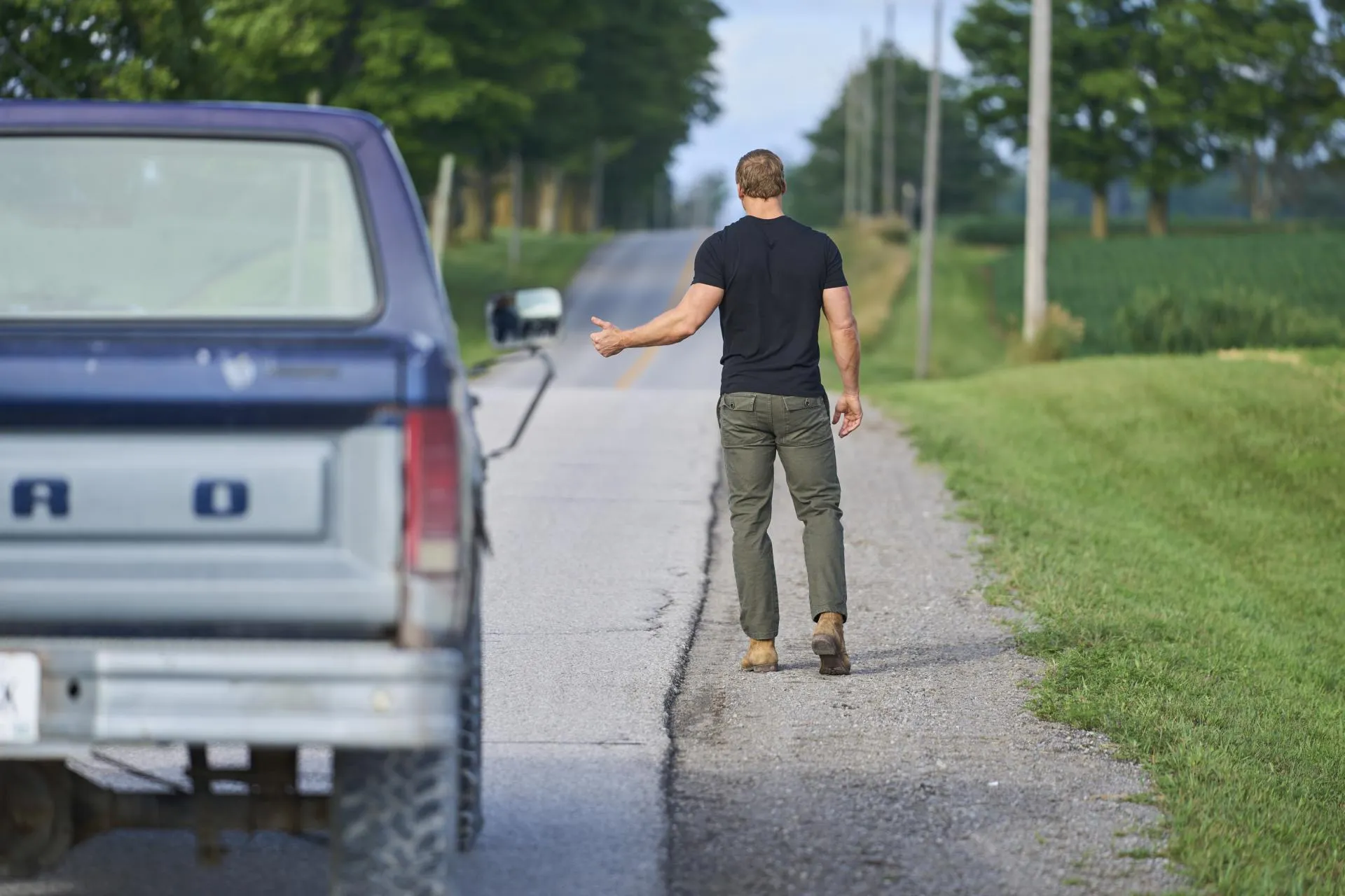 Alan Ritchson as Jack Reacher walking away down a rural roadside with his thumb out to hitch a ride in Reacher season 1