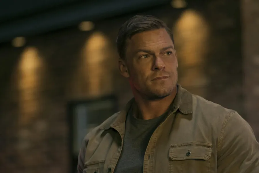 Alan Ritchson as Jack Reacher in a tan work shirt, looking off camera in an alley