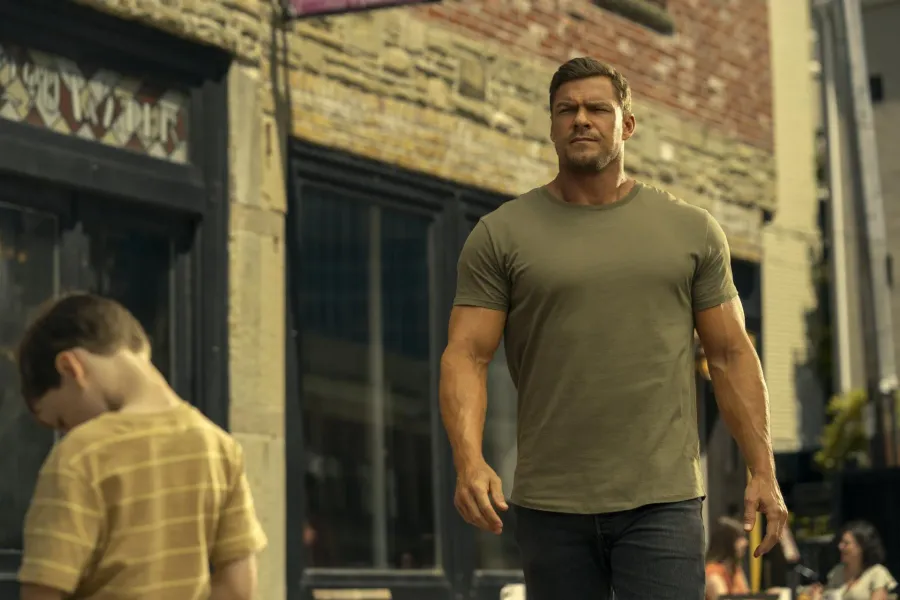 Alan Ritchson as Jack Reacher walking down a small-town street in a fitted olive T-shirt