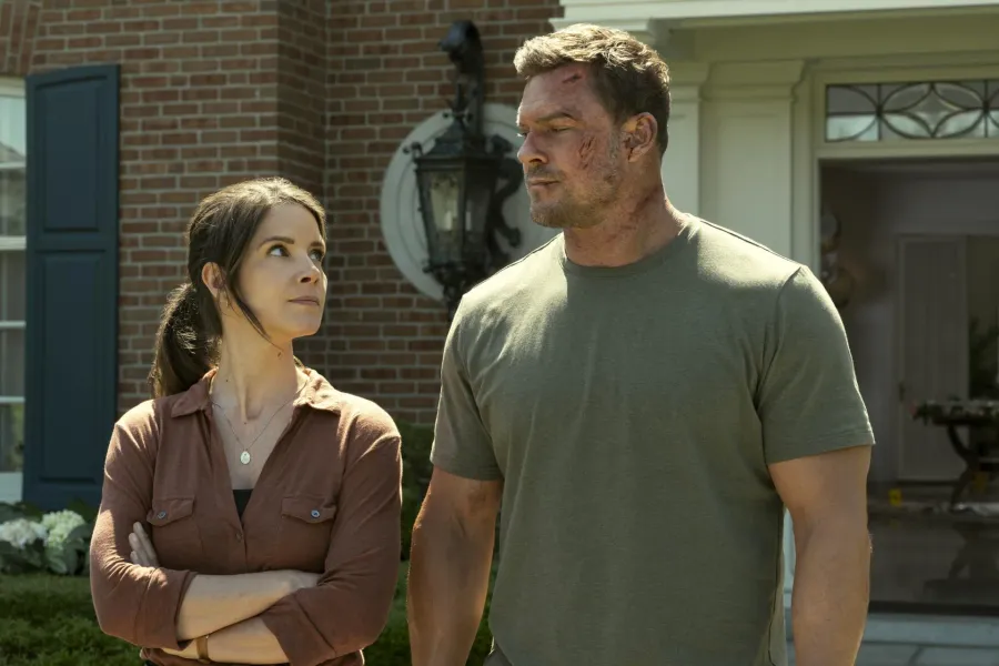 Sonya Cassidy as Susan Duffy standing with a bruised Alan Ritchson as Jack Reacher outside the Beck house