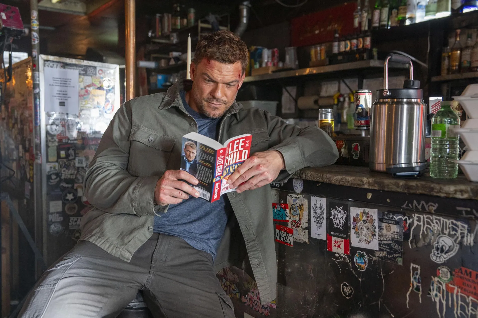 A bruised Alan Ritchson as Jack Reacher reading a Lee Child paperback at a dive bar counter in Reacher season 4