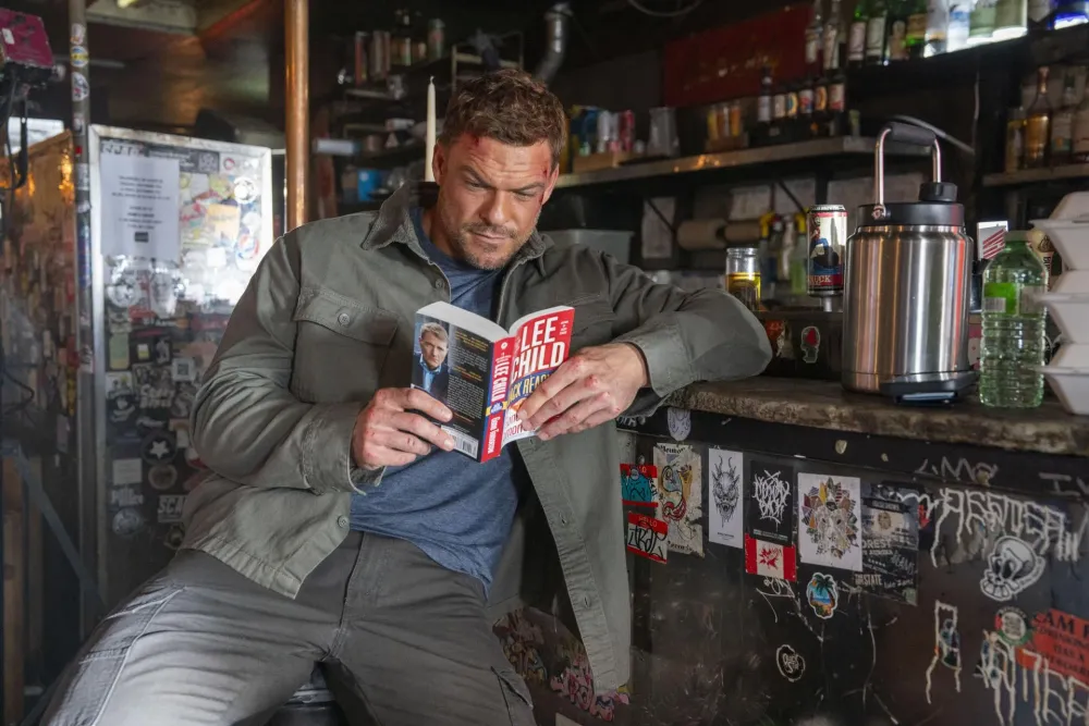 A bruised Alan Ritchson as Jack Reacher reading a Lee Child paperback at a dive bar counter in Reacher season 4